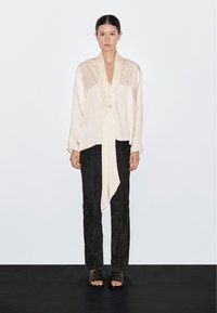 Blusa di seta beige con orlo asimmetrico, davanti drappeggiato e maniche lunghe, abbinata a pantaloni neri aderenti e sandali bassi su una superficie scura.