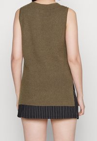 Ärmlös, olivgrön stickad topp med ribbat mönster; sidoförslutningar, lager över ett svart randigt plagg, synlig fålldetalj.