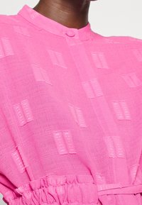 Blusa rosa con texture e colletto a bottoni, caratterizzata da un motivo intrecciato di rettangoli e una vita elastica. Tessuto leggero.