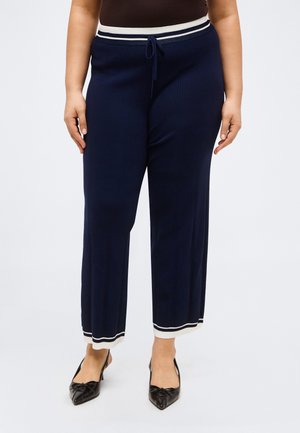Kvinde iført marineblå bukser med vide ben og hvid kant ved talje og kant, kombineret med sorte spidse sko.