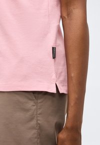 Gros plan d'une personne portant un polo rose clair avec une étiquette noire Peak Performance et un pantalon beige.