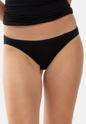 Kobieta ubrana w czarne majtki w stylu bikini oraz czarny krótki top, pokazana od talii do połowy uda na białym tle.