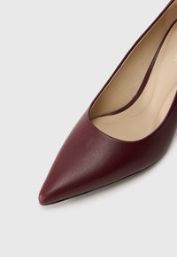 Burgundy nahka korkokenkä, jossa on terävä kärki ja matala V-muotoinen aukko. Sileä ulkopinta ja beige sisävuori.