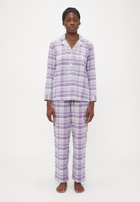 Žena stojí naboso a má na sebe pohodln�ú dlhú nočnú košeľu s gombíkmi v purpurovo-bielej kockovanej vzore a zodpovedajúce nohavice, pričom je na bielom pozadí.
