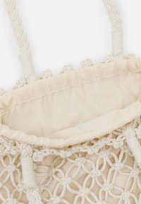 Sac fourre-tout de couleur crème présentant un design tissé et noué, avec des poignées tressées et une doublure intérieure douce et non structurée.