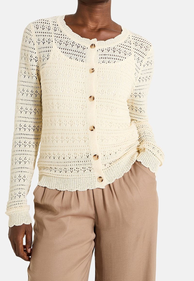 Femme portant un cardigan crème en crochet boutonné avec des bords festonnés et un pantalon ample beige, main dans la poche.