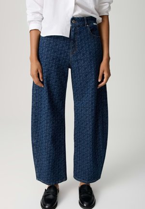 Person trägt dunkelblaue Jeans mit weitem Bein und dezentem Blumenmuster, ein weißes Langarmhemd und schwarze Loafers.