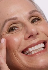 Close-up van een glimlachend gezicht met een gladde huid, lichte make-up en goed onderhouden wenkbrauwen. Opvallende tanden, subtiele rimpels en een hand die de wang aanraakt.