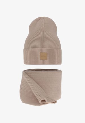 Beige gestrickte Mütze mit umgeschlagenem Bündchen und Etikett, kombiniert mit passendem langen, gestrickten Schal, beide vor weißem Hintergrund präsentiert.