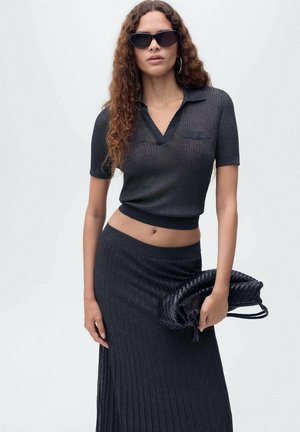 Crni rebrasti pleteni crop top s kratkim rukavima i V-izrezom, uparen s dugom plisiranom suknjom iste boje. Model drži tkanu crnu torbicu.