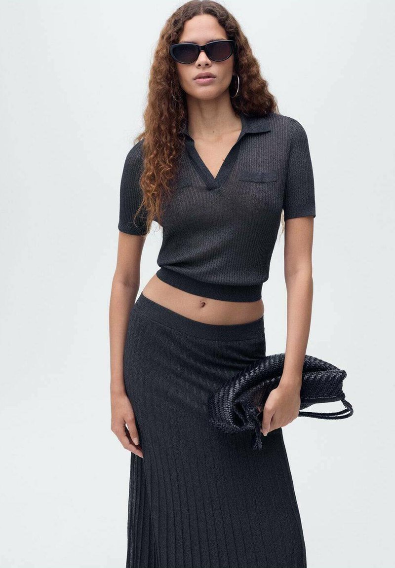 Crni rebrasti pleteni crop top s kratkim rukavima i V-izrezom, uparen s dugom plisiranom suknjom iste boje. Model drži tkanu crnu torbicu.