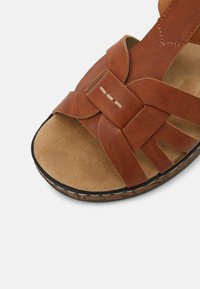 Sandale en cuir marron avec des larges brides, présentant un dessus texturé et une semelle intérieure en suede beige douce. Détails de couture remarquables sur les brides.
