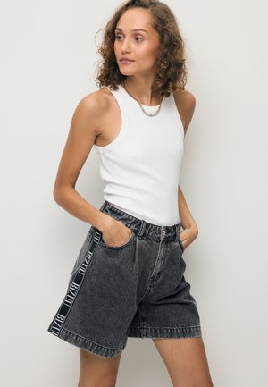 Kobieta w białej bluzce bez rękawów i czarnych, wysokich szortach dżinsowych z oznakowanymi bocznymi paskami, ręce w kieszeniach, patrzy bokiem.