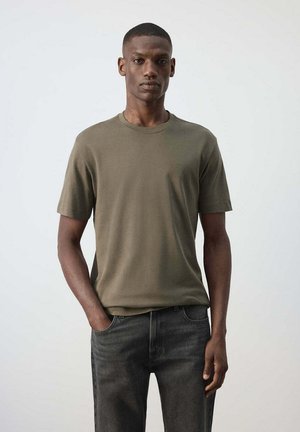 Homme debout devant un fond uni, portant un t-shirt vert olive et un jean gris foncé, une main dans la poche.