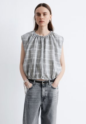 Kvinde iført ærmeløs grå- og hvidternet top med rynket halsudskæring, sort bælte, grå jeans og sølvøreringe, hænderne i lommerne.