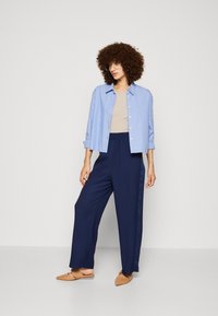 Chemise à rayures bleues et blanches à manches courtes, haut beige, pantalon large bleu marine, et chaussures plates beiges. Tissu lisse, coupe décontractée, design casual.