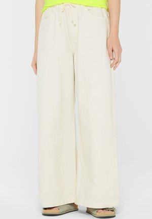 Person wearing wide-leg, high-waist cream pants with drawstring waist and green top, standing with hands relaxed by sides.