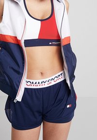 Kobieta w granatowych siatkowych szortach z białym paskiem Tommy Sport, sportowym biustonoszu w czerwono-biało-granatowe wzory oraz dopasowanej kurtce na zamek.