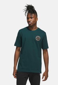Dunkelgrünes Baumwoll-T-Shirt mit kurzen Ärmeln, rundem Halsausschnitt und einem kreisförmigen Grafiklogo in Orange und Schwarz auf der Brust.