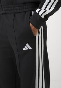 Osoba ubrana w czarne spodnie dresowe i kurtkę Adidas z białym wzorem trzech pasków i logo, z ręką w kieszeni, na jednolitym tle.