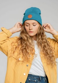 Turkusowy beanie o prążkowanej fakturze z okrągłą brązową skórzaną łatą. W połączeniu z żółtą kurtką z guzikami i białą koszulką.