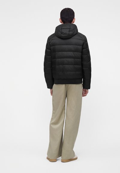 Homme portant une doudoune noire à capuche et un pantalon large beige, debout de dos devant un fond clair uni.