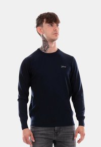 Maglione blu navy a maniche lunghe con scollatura rotonda e polsini a coste, caratterizzato da un piccolo logo ricamato sul petto. Texture liscia.