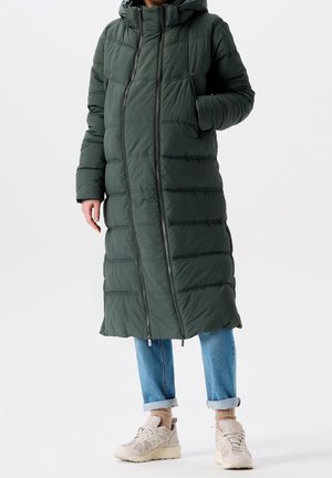 Persoon die een donkergroene, lange gewatteerde jas met capuchon draagt, blauwe opgerolde spijkerbroek, beige sokken en beige sneakers, staand tegen een witte achtergrond.