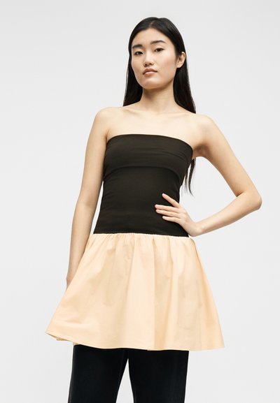 Νεαρή γυναίκα με μακριά μαύρα μαλλιά, φοράει μαύρο τοπ χωρίς τιράντες με επικάλυψη από μπεζ φουσκωτή φούστα και μαύρο παντελόνι, στέκεται με το χέρι στη μέση.