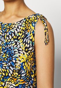 Top sin mangas con estampado floral en negro, que presenta patrones en amarillo, azul y blanco. Tirantes decorativos con lazos y cuentas en los hombros.