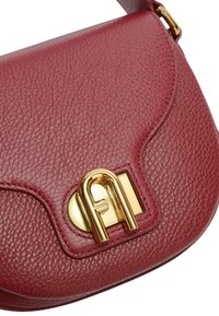 Červená texturovaná kožená kabelka s zakřiveným klopou a zlatým kovovým uzávěrem ve tvaru propletených písmen.