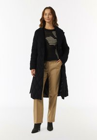 Black quilted long coat with a notched collar, worn over a dark striped sweater, paired with beige trousers and black ankle boots.
