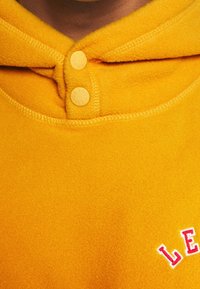 Sudadera con capucha de forro polar amarillo con textura suave, que cuenta con dos botones beige en el cuello y letras rojas en el frente.