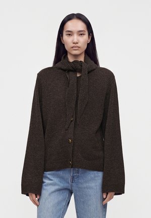 Vrouw met stijl zwart haar, die een donkerbruine, gesloten cardigan draagt met een gestrikt sjaaldetail en lichtblauwe spijkerbroek, staand tegen een effen achtergrond.