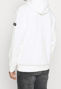 Vit huvtröja med mjuk textur, som har en logotyp på vänster ärm och en avslappnad passform. I kombination med mörka jeans.