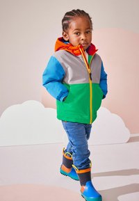 Chaqueta colorida con paneles grises, verdes, azules y naranjas, cierre de cremallera y dos bolsillos. Lleva puestas unos vaqueros azules y unas botas de goma multicolores.