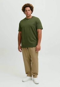 Homem com cabelo encaracolado, vestindo uma t-shirt verde-oliva, calças bege, ténis brancos e um colar fino, em pé sobre um fundo claro e simples.
