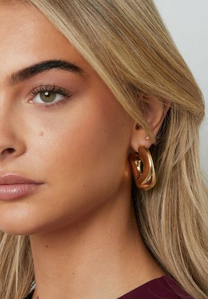 Nahaufnahme eines Frauenkopfes mit blonden Haaren, grünem Auge und großem, verdrehtem goldfarbenem Creolen-Ohrring am sichtbaren Ohr.