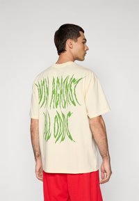 Mand iført en cremefarvet t-shirt med stor forvrænget grøn tekst på ryggen og løse røde bukser, som står op ad en ensfarvet lys baggrund.