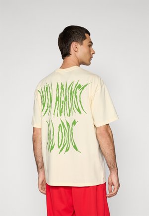 Mand iført en cremefarvet t-shirt med stor forvrænget grøn tekst på ryggen og løse røde bukser, som står op ad en ensfarvet lys baggrund.