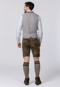 Grau gestreiftes Westchen mit Rückenversteller, getragen über einem hellen Hemd. Braune Lederhosen und graue gestrickte Beinstulpen. Dunkle Schuhe vervollständigen das Outfit.