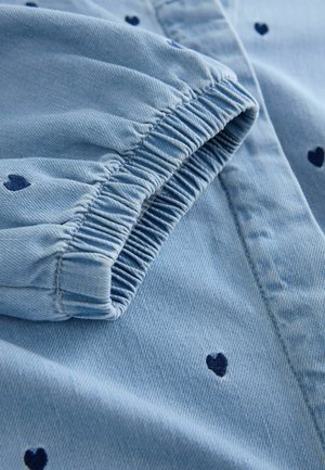 Šviesiai mėlyna džinsinė marškinėliai su mėlynais širdies raštais, elastingu rankogaliu ir tekstūruota medžiaga. Artimas vaizdas į detales.