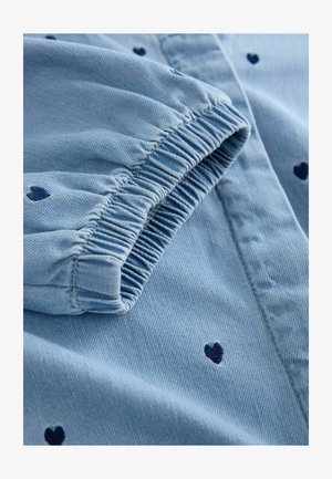 Jasnoniebieska koszula dżinsowa z niebieskimi wzorami serc, z elastycznym mankietem na rękawie i teksturowaną tkaniną. Zbliżenie na detale.