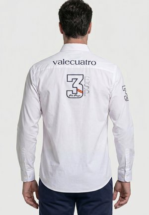 Hombre vestido con camiseta blanca de manga larga con el texto "valecuatro" en la parte superior de la espalda y el logo "3 ESP" con detalles de la bandera en el centro de la espalda.