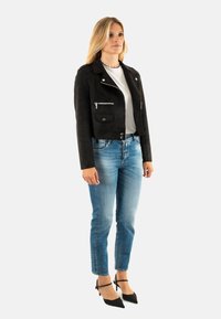 Chaqueta corta de ante negra con cremallera y bolsillos con solapa, combinada con jeans azul claro y tacones negros de punta. Camiseta gris sencilla debajo.