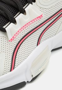 Zapatilla con parte superior de malla en gris claro, que presenta cordones negros, costuras rosas y una suela blanca con acentos negros. Detalle transparente en el talón.