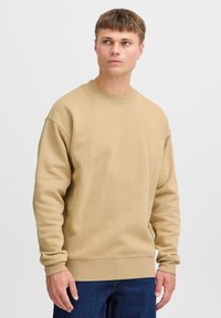 Beige kapuutsiga särgid ümmarguse kaelusega ja soonikkoes mansettidega. Omab pehme tekstuuri ja mugavat lõiget. Kanda tumedate teksadega.
