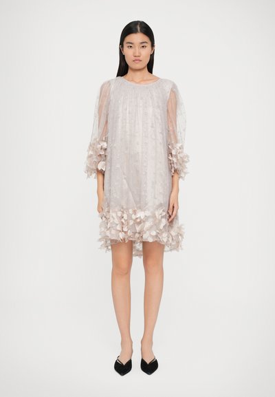 Vestido ligeiramente rosa em tecido transparente, com embelezamentos florais na bainha e mangas. Decote redondo, corte solto e uma textura delicada. Sapatos pretos de bico fino.