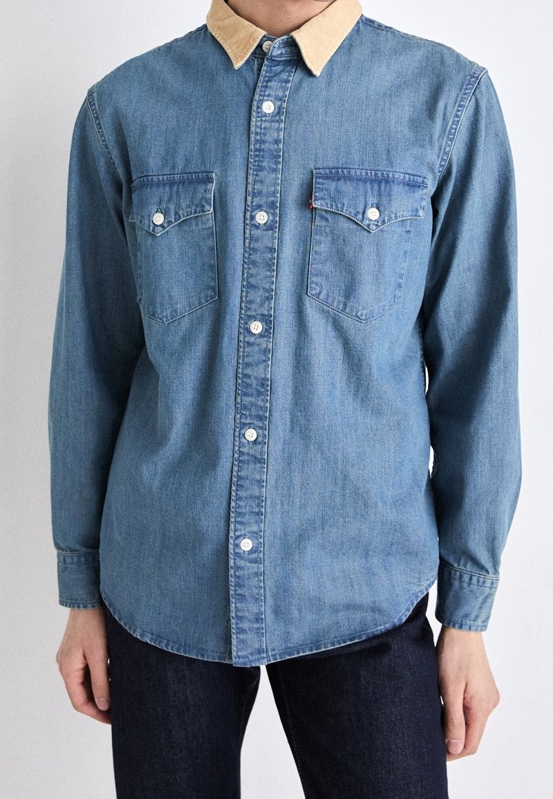 Mann trägt ein blaues Jeanshemd mit beigem Kragen und geknöpften Brusttaschen, kombiniert mit dunkler Hose, steht vor einem schlichten Hintergrund.