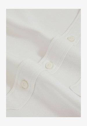Fehér textúrájú szövet, amely finom bordázott mintázattal rendelkezik, a gombolás mentén pedig több kis, kerek fehér gomb található.
